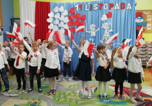 Dzieci w eleganckich strojach machają flagami w rytm muzyki.