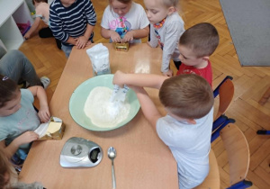 Dodawanie cukru pudru przez chłopca, a dookoła stolika siedzą dzieci.