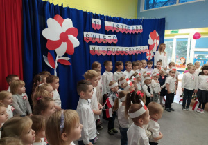 Starszaki podczas mówienia wiersza na tle niebieskiej dekoracji z biało-czerwonymi akcentami.