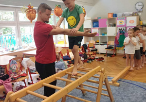 Chłopiec idący po drabinkach w asekuracji nauczyciela.
