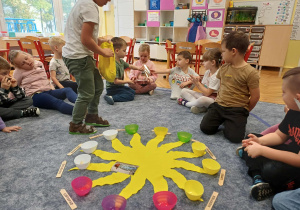Chłopiec chodzący dookoła słoneczka wrzucający koraliki, jako symbol miesiąca, w tle dzieci przekazujące sobie zdjęcie jubilata z tego okresu.
