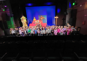 Zdjęcie dzieci z grupy Jagódki i Krasnoludki przedsceną, na której stoją aktorzy z lalkami.