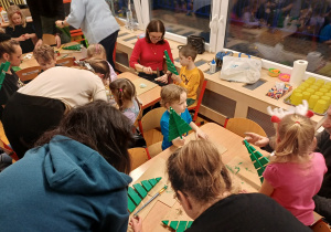 Grupa dzieci i zaproszonych gości podczas pracy, na różnym etapie wykonania ozdoby bożonarodzeniowej.