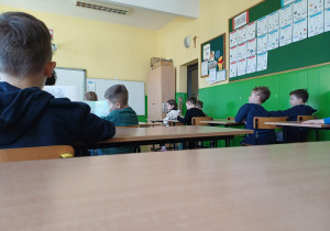 Dzieci słuchające nauczycielki siedzą w ławkach pod ścianą.